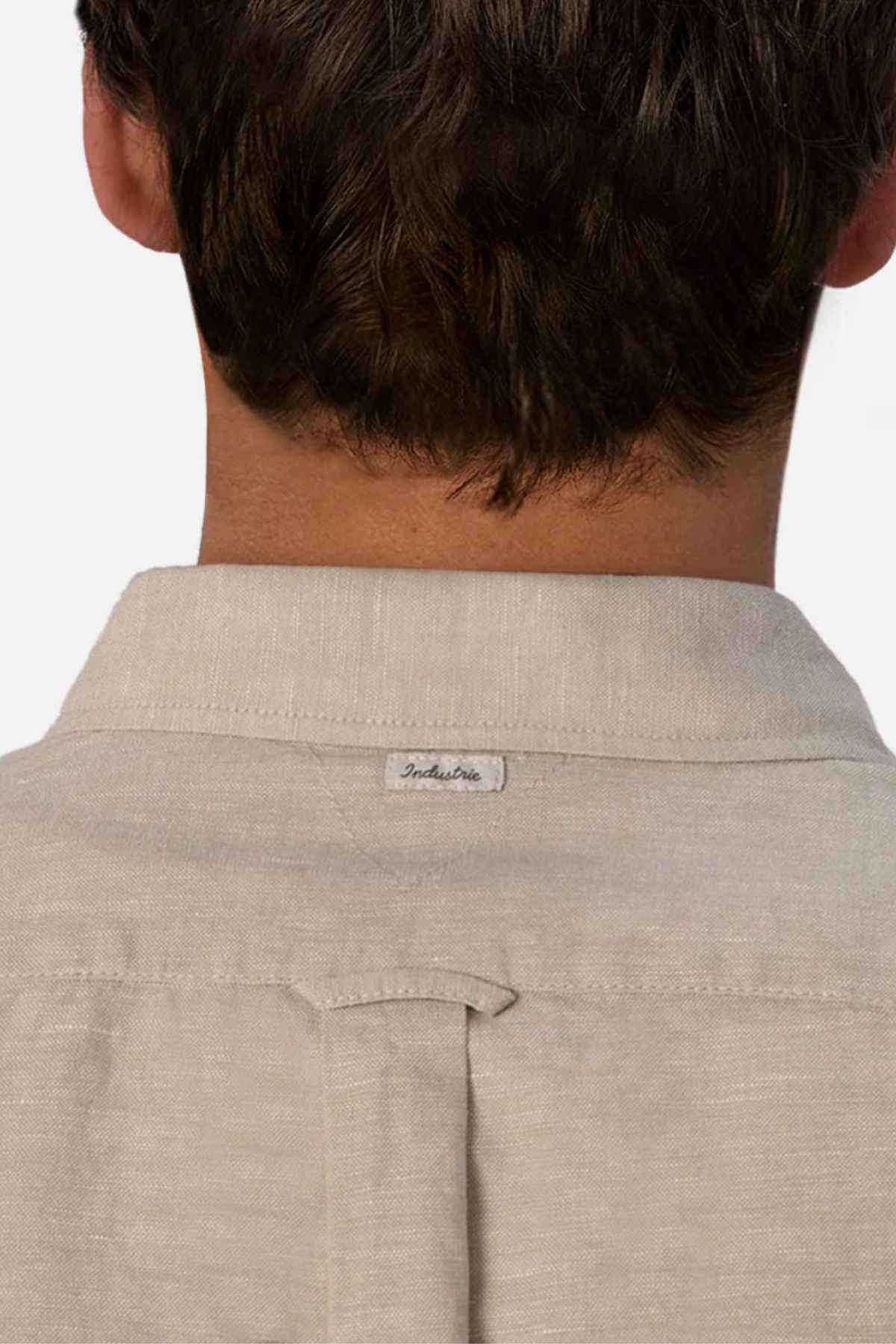 A person is seen from the back wearing the Industrie The Tennyson Linen Blend Shirt in beige. The spread collar and a small tag labeled "Industrie" are visible, with their head turned away from the camera, embodying a casual summertime style.