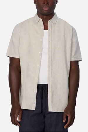 A person wearing the Industrie The Tennyson Linen Blend Short Sleeve Shirt in light beige over a white tank top, paired with dark pants, stands against a plain white background, exuding summertime comfort.