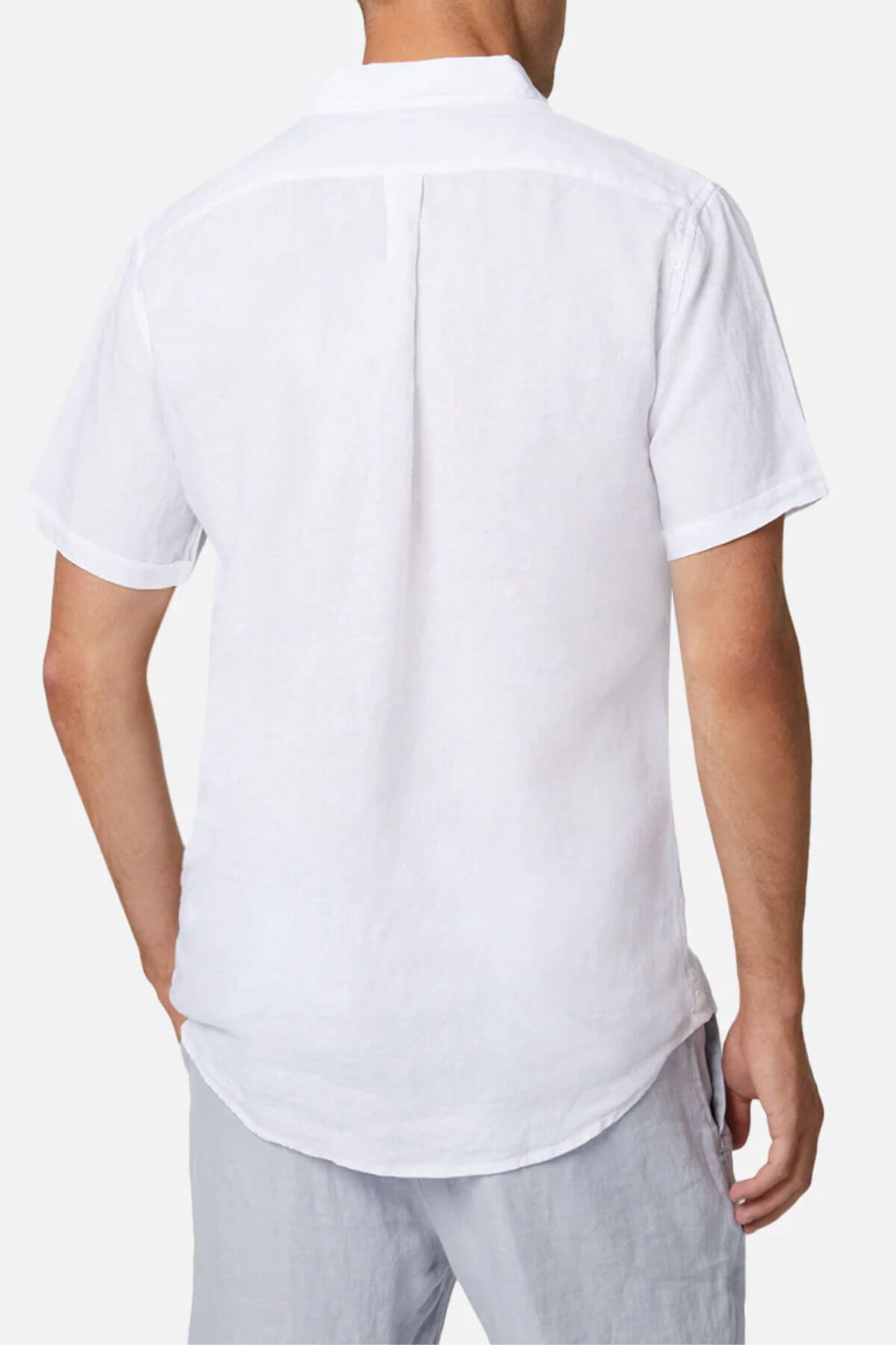 Someone is wearing an Industrie The Tennyson Linen Short Sleeve Shirt, a white button-up with a smooth texture, paired with light gray pants. The setting features a plain white backdrop, and the outfit is viewed from behind in a casual style.