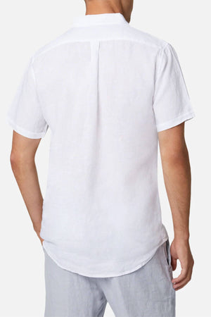 Someone is wearing an Industrie The Tennyson Linen Short Sleeve Shirt, a white button-up with a smooth texture, paired with light gray pants. The setting features a plain white backdrop, and the outfit is viewed from behind in a casual style.