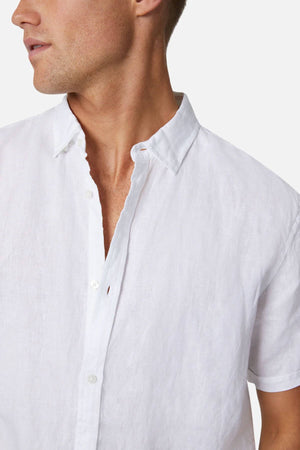 A man wears the Industrie The Tennyson Linen Short Sleeve Shirt in a casual white, capturing a relaxed vibe with its slightly wrinkled fabric. He is shown from shoulders to chest, facing slightly left against a plain backdrop.