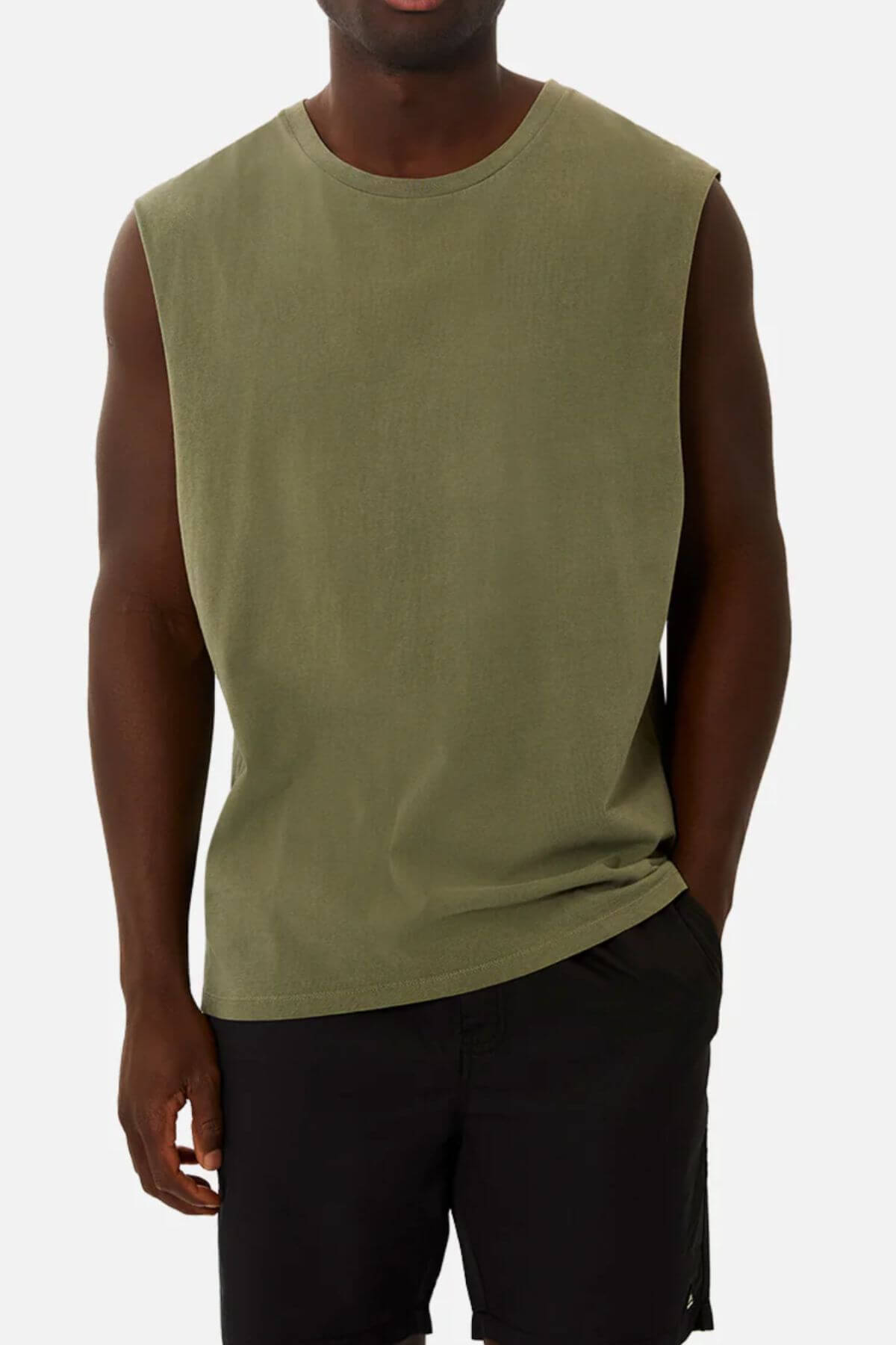 A person in an olive green Industrie The Triple G Sleeveless Tee, made from 100% cotton, and black pants poses against a white backdrop. Their right hand is casually in their pocket, and the image captures them from shoulders to thighs.