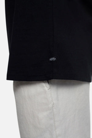 Close-up of a person wearing the Industrie The Triple G Sleeveless Tee in black, showcasing a relaxed fit with a small hem label, paired with light-colored pants. Focus is on the lower half of the shirt and the top section of the pants.