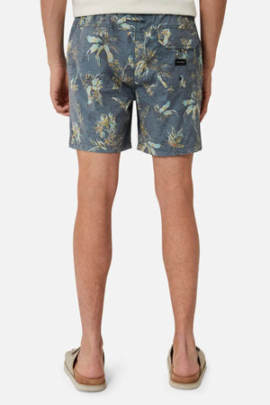 A man faces away from the camera, wearing Industrie The Viloria Bahama Short—blue floral-patterned swim shorts with an elasticised waistband—and beige flip-flops, set against a plain white background.