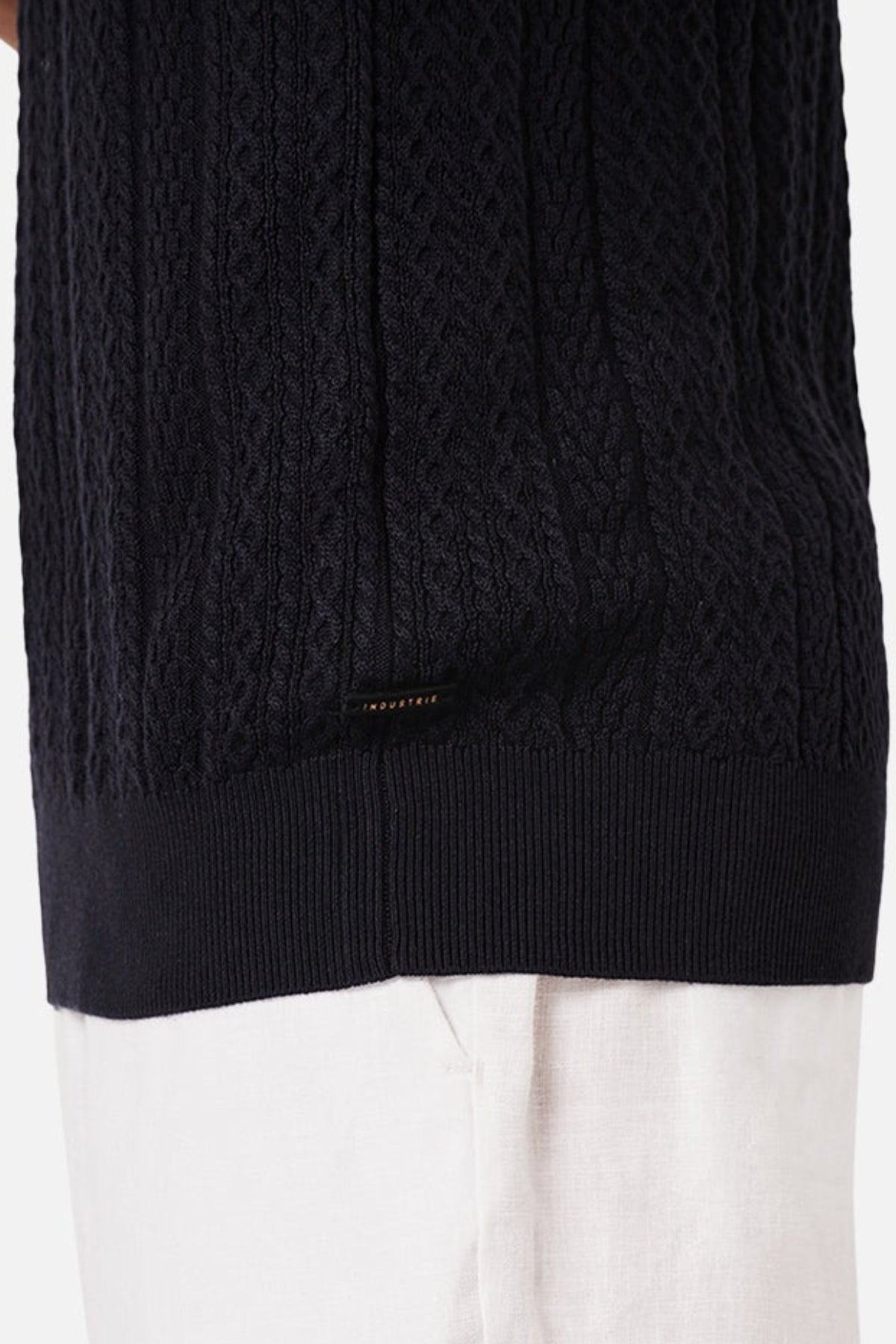 A person wears Industrie The Vito Polo, a black cable-knit sweater, with white pants. The detailed knit is enhanced by a small black label at the bottom, emphasizing texture and garment contrast.