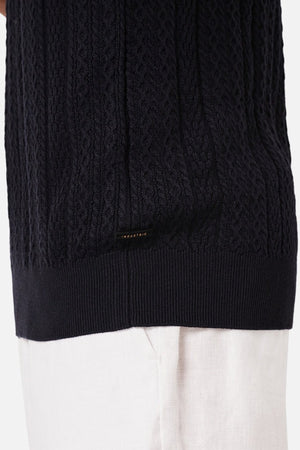 A person wears Industrie The Vito Polo, a black cable-knit sweater, with white pants. The detailed knit is enhanced by a small black label at the bottom, emphasizing texture and garment contrast.