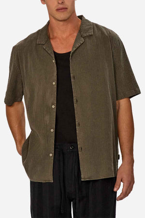 A person wearing a short-sleeve textured olive green Industrie The Washed Magna Short Sleeve Shirt layered over a black tank top and black pants, with hands partially in pockets. The breathable fabric ensures comfort against the plain white backdrop.