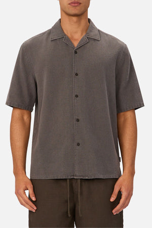A man wears the Industrie The Washed Menorca Short Sleeve Shirt, a charcoal gray cotton blend camp collar shirt with a relaxed fit, styled with dark pants and his arms at his sides.