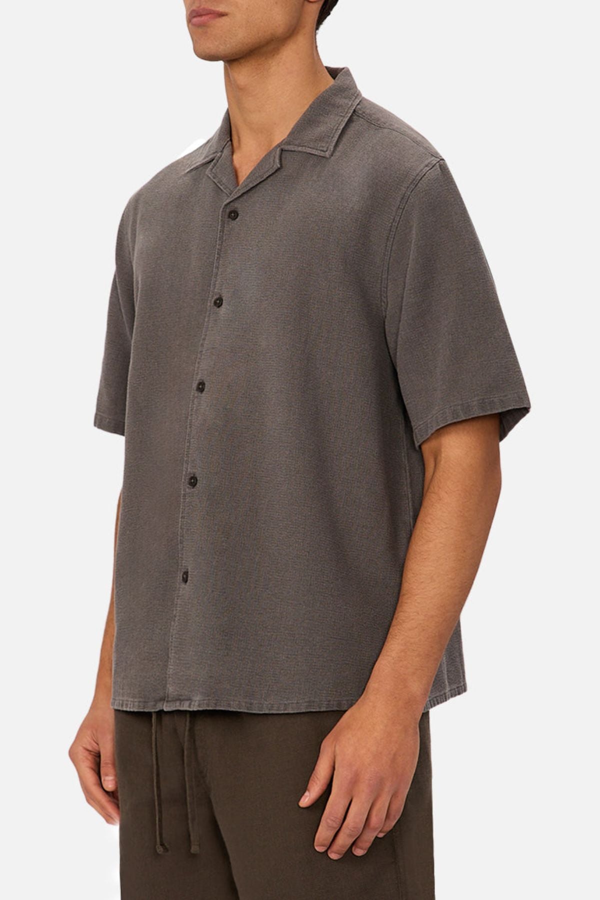 A man wears the Industrie The Washed Menorca Short Sleeve Shirt in gray with dark brown pants, standing against a plain white background. His face is partially out of frame.