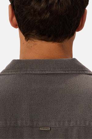 A close-up of the back of a person’s head and shoulders, highlighting short dark hair and an Industrie The Washed Menorca Short Sleeve Shirt in textured gray with a camp collar and a small rectangular tag near the collar.