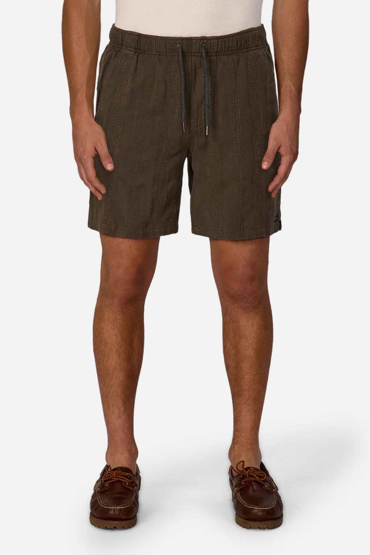 A person dressed in Industrie The Washed Salamanca Short in dark green, created from a textured cotton blend, pairs them with brown leather shoes against a plain white background. The shorts display a subtle pattern and tailored fit, while the shoes feature visible stitching and laces for additional style.