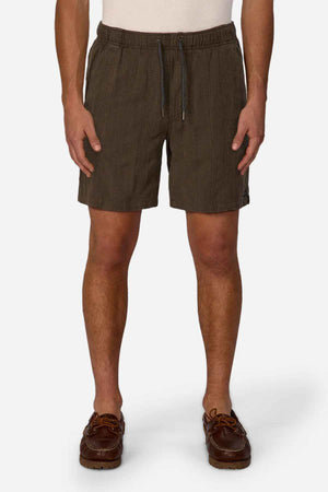 A person dressed in Industrie The Washed Salamanca Short in dark green, created from a textured cotton blend, pairs them with brown leather shoes against a plain white background. The shorts display a subtle pattern and tailored fit, while the shoes feature visible stitching and laces for additional style.
