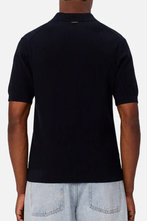 A person wearing the Industrie The Valencia Polo, a short-sleeve black 100% cotton shirt, with light blue jeans, shown from the back against a plain light background.