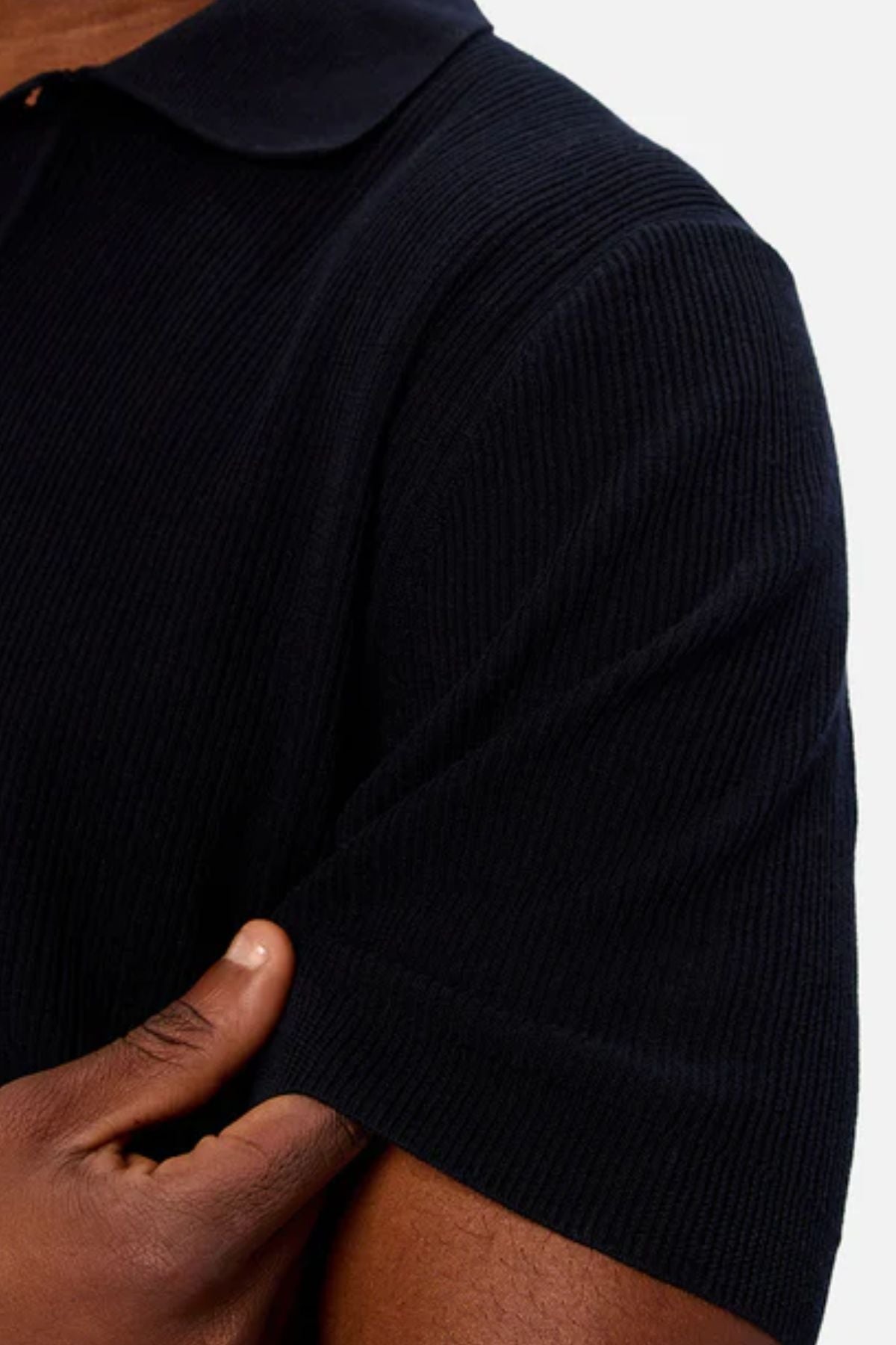 Close-up of a person’s arm and torso wearing the Industrie The Valencia Polo, a short-sleeved black shirt with ribbed texture made from breathable fabric. Their left hand rests on their right arm against a plain light background.