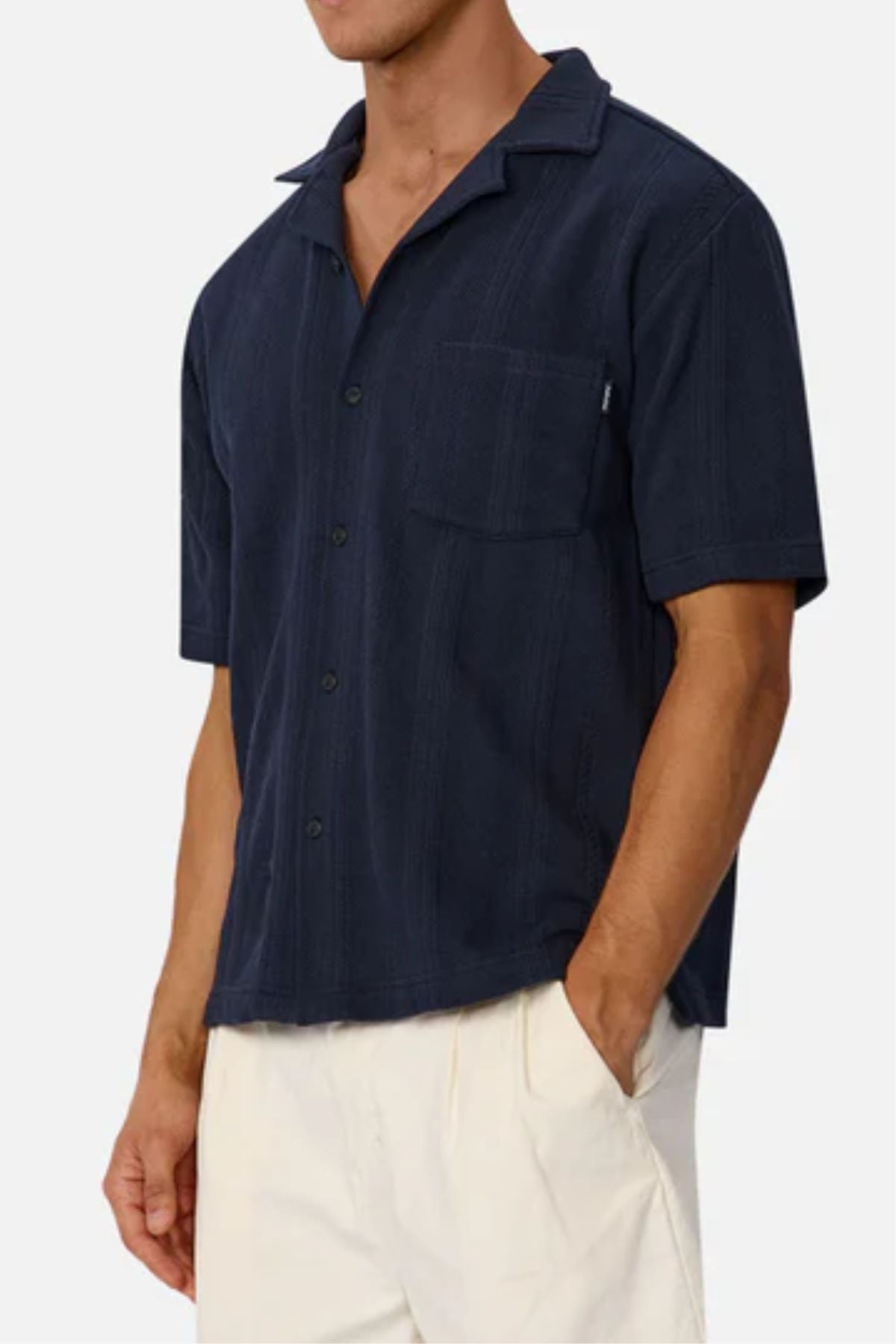A man wears the Industrie The Malaga Short Sleeve Shirt, a navy camp collar top with chest pocket, paired with off-white pants. His face is unseen as he stands with his left hand in his pocket.