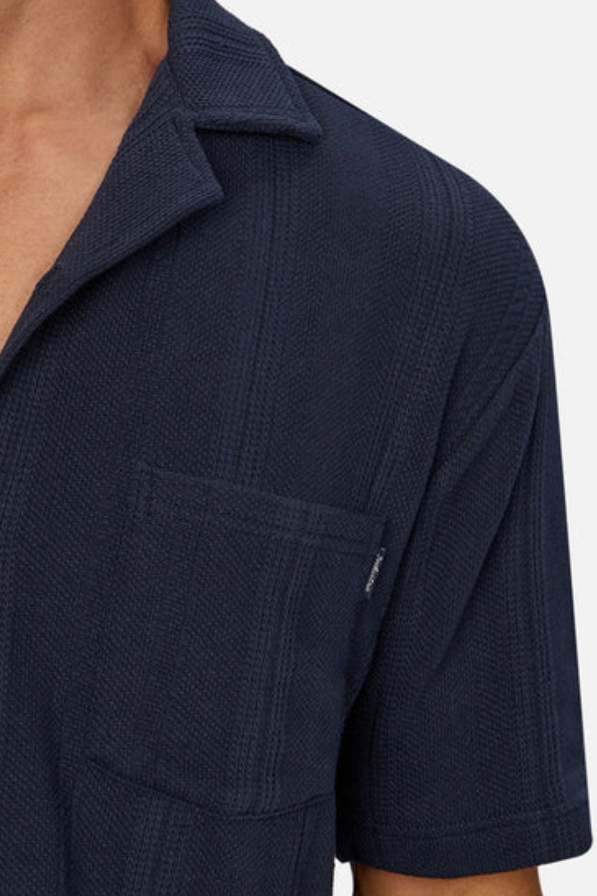 A close-up shows a person wearing the Industrie The Malaga Short Sleeve Shirt in textured dark navy, featuring a camp collar, chest pocket, and subtle vertical stripes. The neck and collarbone are partially visible.