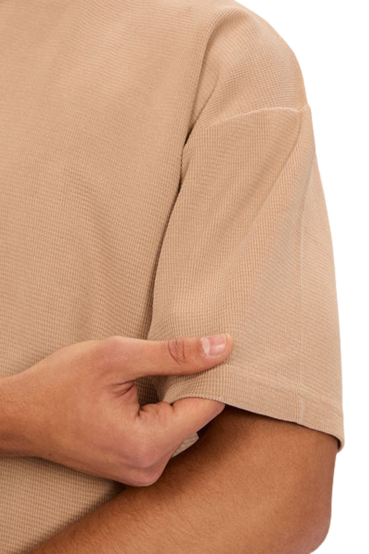 Close-up of a person wearing the Industrie The Marlon Tee, a beige short-sleeved waffle knit shirt, holding their sleeve with their thumb and fingers against a white background.