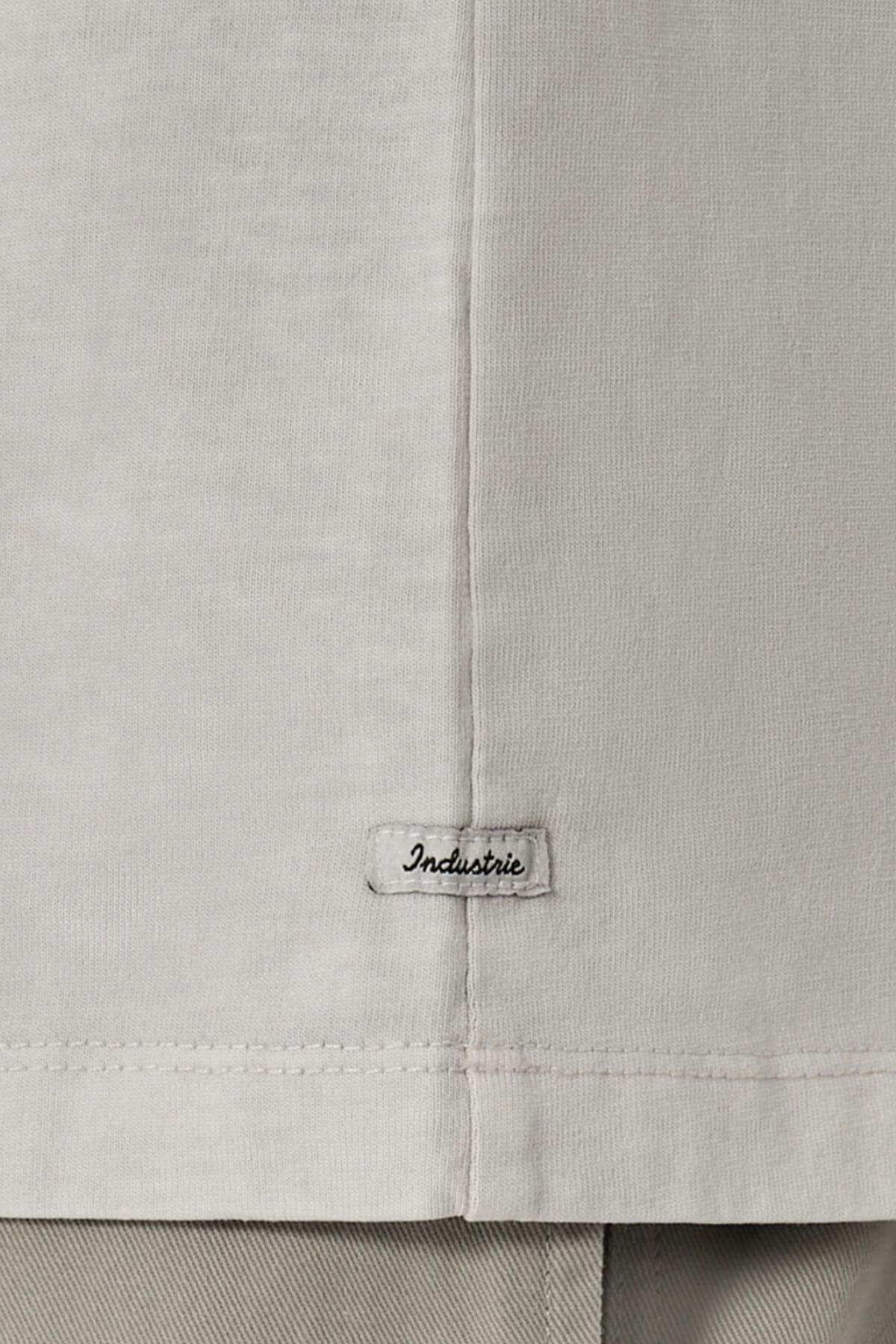 Close-up of the lower part of the Industrie The Del Sur Tee in light grey, featuring an oversized fit and a small stitched "Industrie" label near the hem. Grey pants are visible below this 100% cotton shirt.