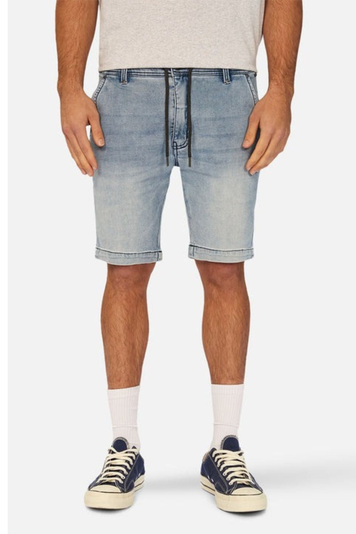 A person wearing Industrie The Drifter Denim NC Short, which is a slim fit light denim short with a cuffed hem and a black drawstring, paired with a grey shirt, white socks, and navy canvas sneakers, stands against a plain background. The image shows the person from the shoulders down.