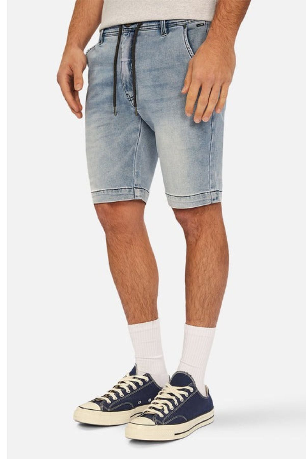 A person wearing the Industrie The Drifter Denim NC Short in light blue, featuring a black drawstring and cuffed hem, complemented by white socks and slim-fit navy blue sneakers with white soles against a plain white background.