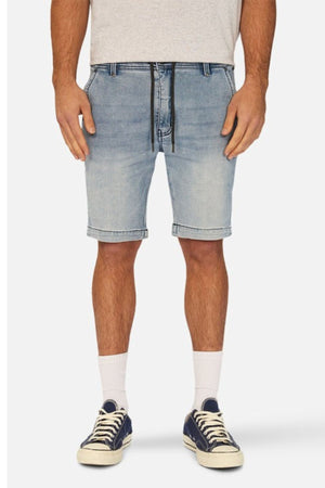 A person wearing Industrie The Drifter Denim NC Short, which is a slim fit light denim short with a cuffed hem and a black drawstring, paired with a grey shirt, white socks, and navy canvas sneakers, stands against a plain background. The image shows the person from the shoulders down.