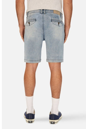 A person wearing a light gray t-shirt, the Industrie The Drifter Denim NC Short in faded blue with a cuffed hem, white knee-length socks, and black sneakers with white soles stands with their back facing the camera.