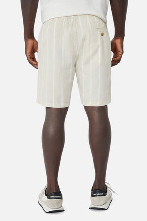 A person effortlessly displays their casual wardrobe in beige and white striped Industrie The Bel Air Shorts, featuring a back pocket button. They pair this with white New Balance sneakers, creating an ensemble that exudes laid-back style against a plain light background.