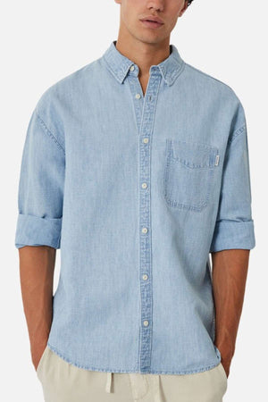 A man wearing Industrie's The McKinley Long Sleeve Shirt, a light blue lightweight denim button-up with a collar, front chest pocket, and sleeves casually rolled up to the elbows. He is standing against a plain white background, with the shirt slightly untucked over beige pants.
