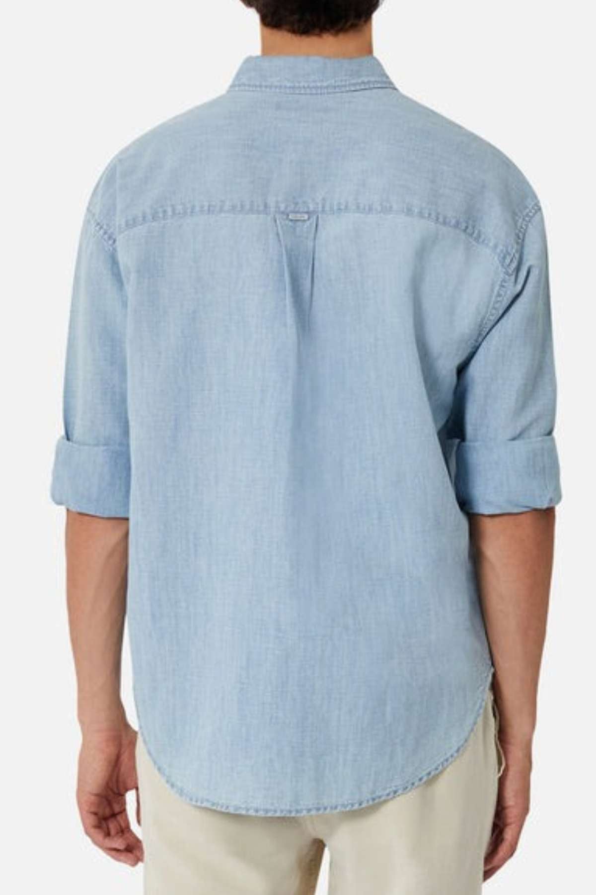A person with rolled-up sleeves is seen from the back, wearing a lightweight denim Industrie The McKinley Long Sleeve Shirt. The shirt features a center pleat and is untucked over beige pants, with subtle INDUSTRIE branding visible. The background is plain white.