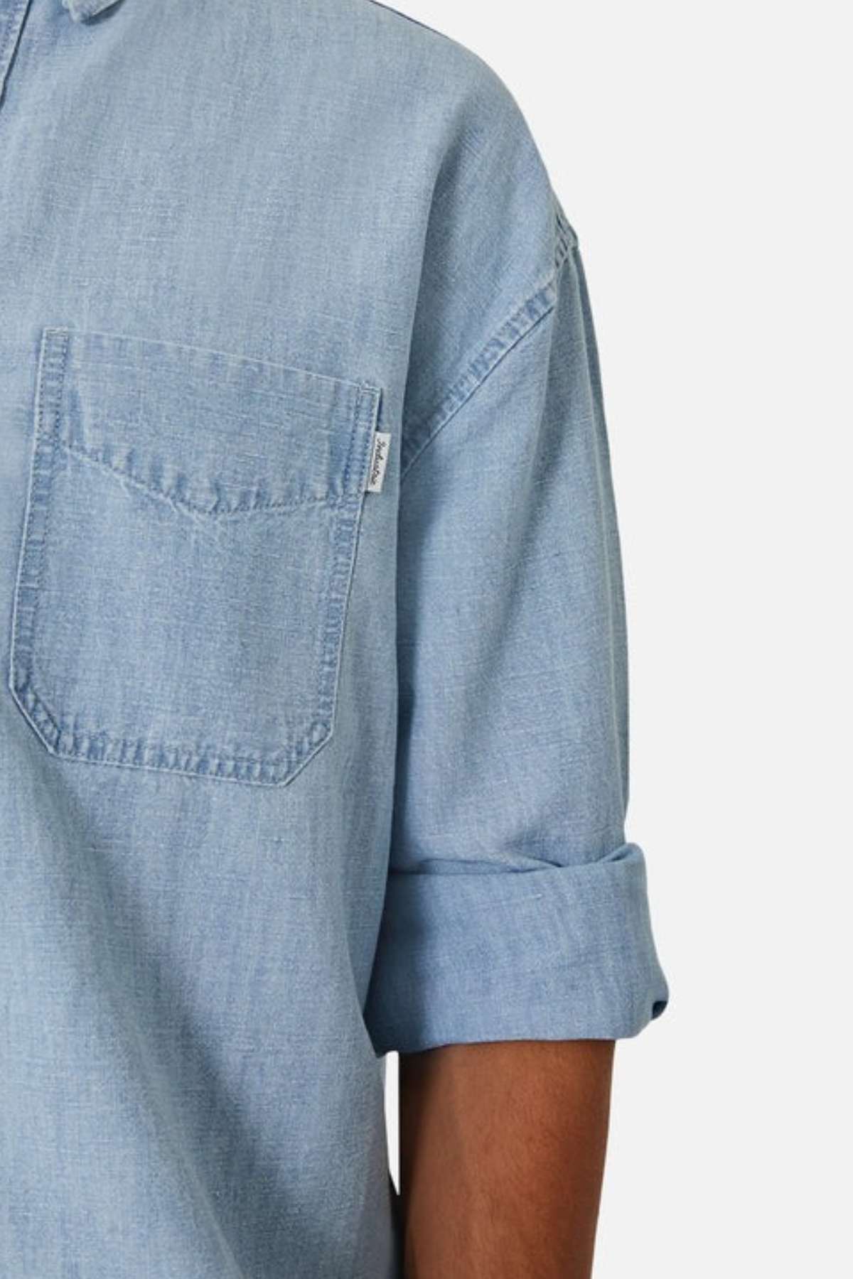 Close-up of a person wearing Industrie The McKinley Long Sleeve Shirt in light blue lightweight denim, showcasing its chest pocket on the left side. This stylish shirt features rolled-up sleeves and INDUSTRIE branding on a small tag on the pocket, set against a plain white background.