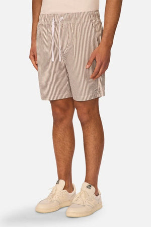 Someone wearing the Industrie The New Southwest Short in cream and brown vertical stripes with a white drawstring, paired with off-white sneakers, stands against a plain light background.