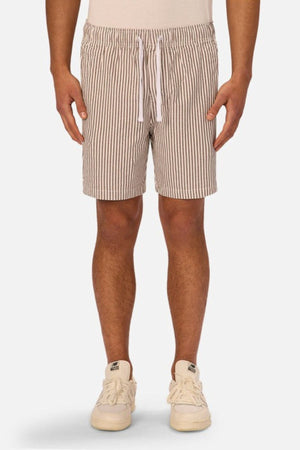 A person wears beige and white vertical striped Industrie The New Southwest Short linen shorts and white sneakers, standing against a plain light background. The image is cropped from the waist down to highlight the classic style of the shorts.