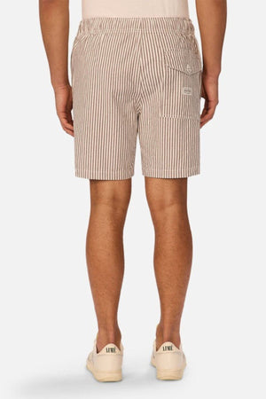 A person stands facing away from the camera wearing Industrie The New Southwest Short men's linen shorts in brown-and-white vertical stripes with a back buttoned pocket and a side cargo pocket, paired with beige sneakers. The background is plain light gray.