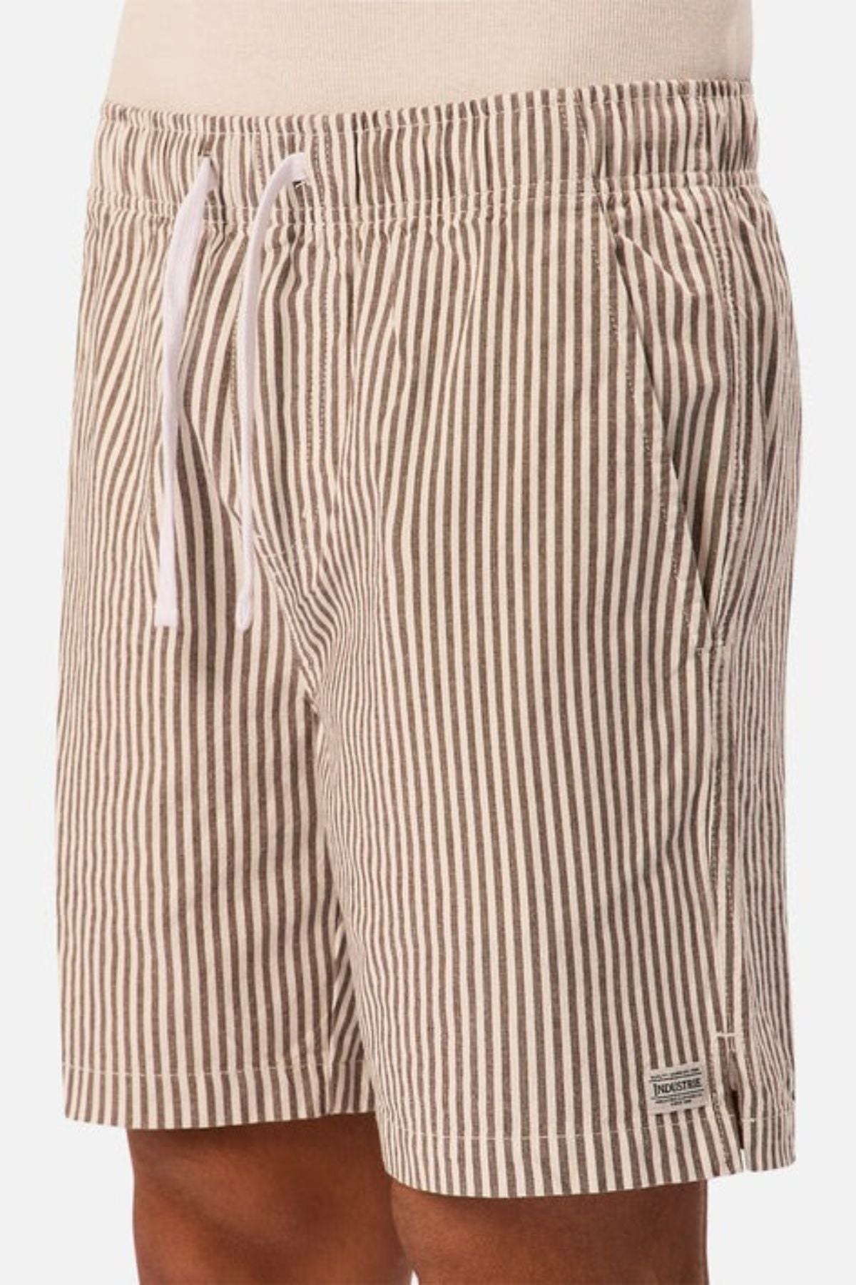 A person wears beige and white vertical striped linen men’s shorts with an elastic waistband, white drawstring, side pockets, and a small rectangular label near the hem: Industrie The New Southwest Short.