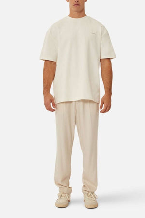 A man faces forward in Industrie's The Newport Tee, highlighting its oversized fit and cream hue, paired with matching pants and beige sneakers. The heavyweight fabric drapes elegantly against a plain white background.