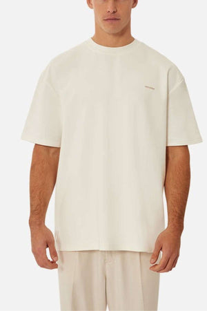 Against a plain background, a person wears Industrieβs The Newport Tee, made of heavyweight fabric. It has an oversized fit and short sleeves with minimal design. Their hands rest by their sides, capturing the relaxed vibe of this monochromatic outfit.