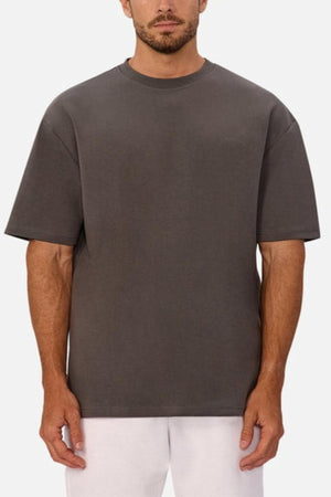 A man wearing a loose-fitting, short-sleeved Industrie The Newport Tee in heavyweight fabric and white pants stands facing forward against a plain light background.
