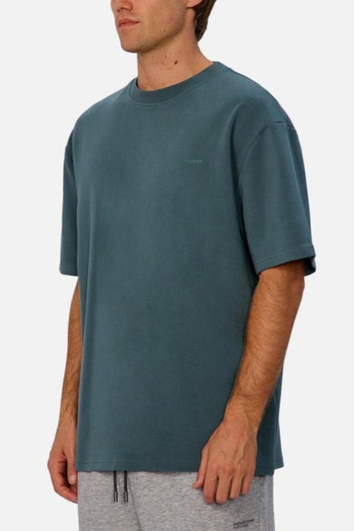 A man stands against a plain light background, wearing Industrie The Newport Tee in oversized teal and light gray sweatpants, with his arms relaxed by his sides.