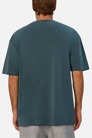 A person with light brown hair is shown from behind, wearing the Industrie The Newport Tee in teal and light gray shorts—ideal for a casual wardrobe—against a plain light background.