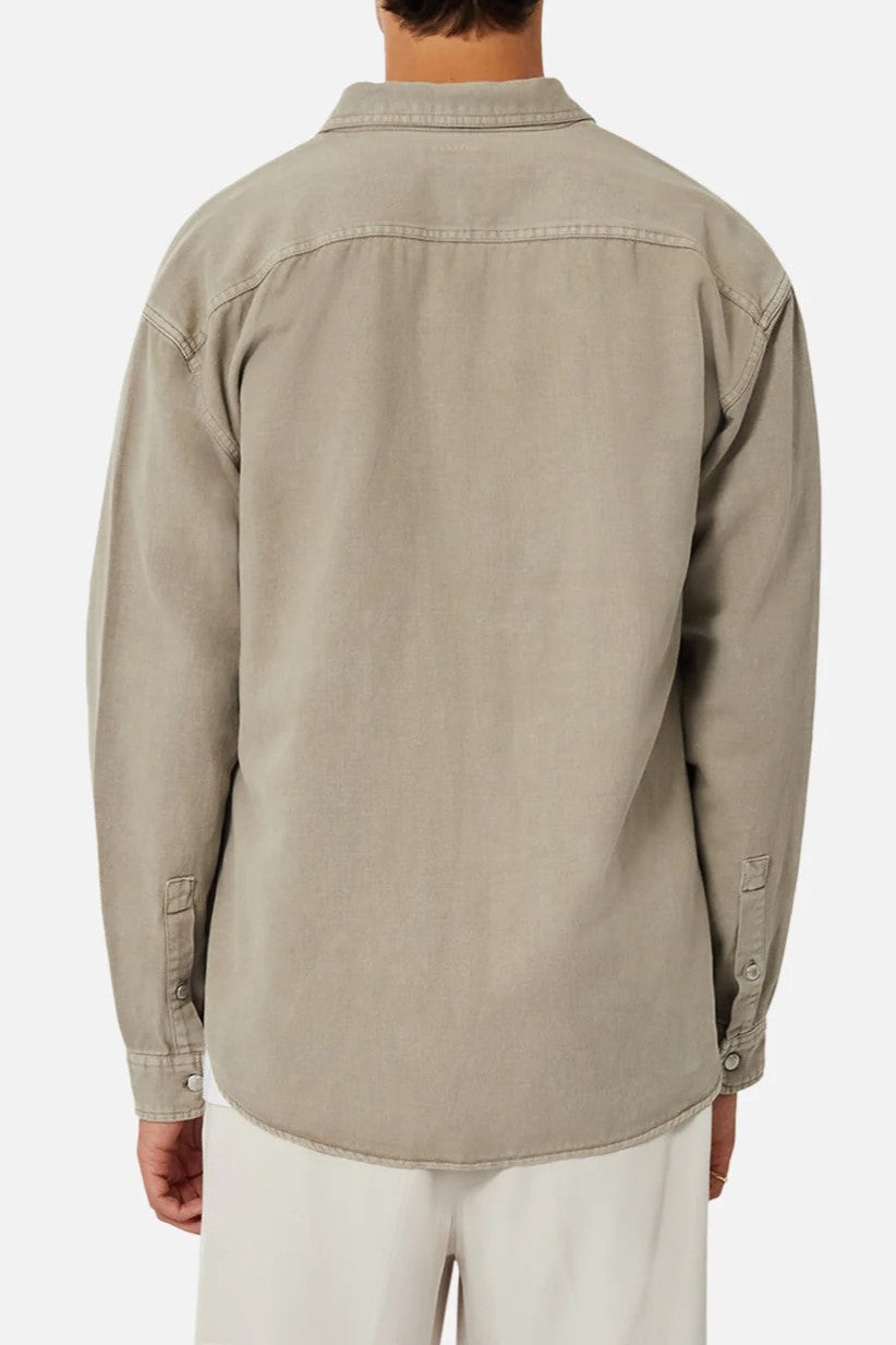 A person is seen from the back wearing Industrie The Portland Shirt, a relaxed-fit light gray denim jacket with buttoned cuffs, resembling an overshirt. It’s paired with cream-colored trousers for a casual look against a plain background.