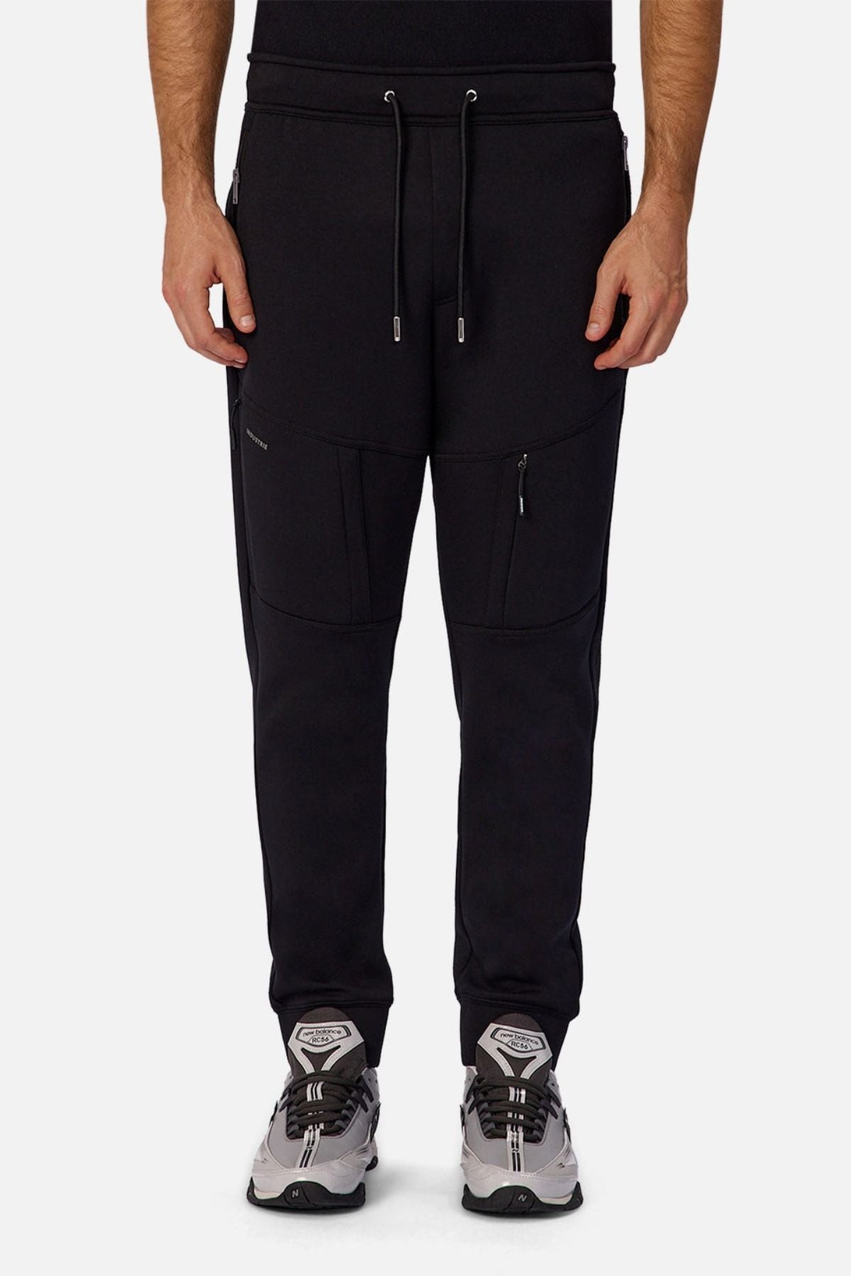 A person wears Industrie The Tech Armoured Track Pant, featuring a drawstring waist and combat pockets, paired with gray and white sneakers. Only the lower half of the body is visible against a plain background.