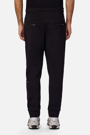 A person wearing Industrie The Tech Armoured Track Pant joggers with zippered back pockets and white sneakers is shown from behind against a plain light background, highlighting the pants' tactical design.