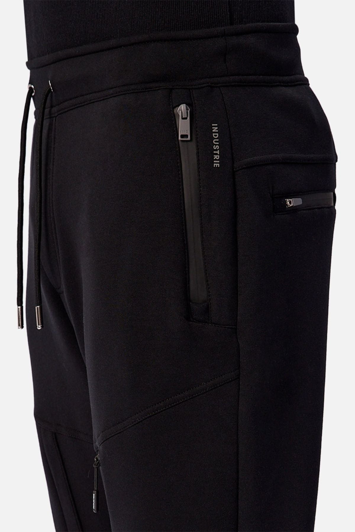 Close-up of a person wearing Industrie The Tech Armoured Track Pant in black, featuring zippered combat pockets, drawstrings, modern seams, and a small INDUSTRIE label by the pocket for a sleek, tactical style.