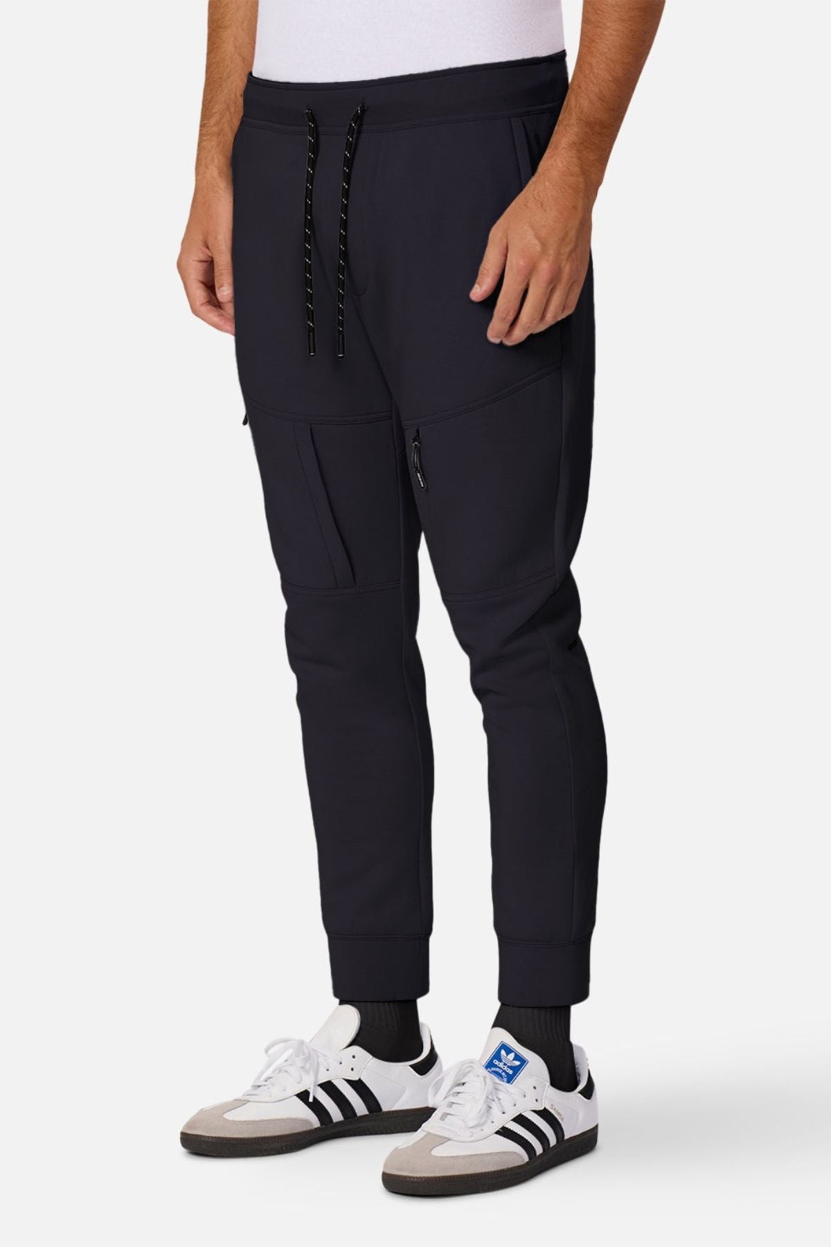 A person wearing Industrie The Tech Armoured Track Pant joggers with ankle cuffs and combat pockets, a white shirt, black socks, and white Adidas sneakers with black stripes stands against a plain white background.