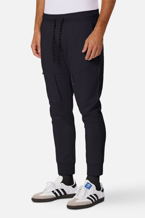 A person wearing Industrie The Tech Armoured Track Pant joggers with ankle cuffs and combat pockets, a white shirt, black socks, and white Adidas sneakers with black stripes stands against a plain white background.