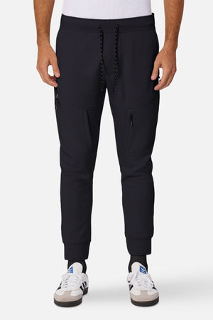 A person wears Industrie The Tech Armoured Track Pant in black with combat pockets and white sneakers, standing against a plain white background. Only the body from the shoulders down is visible.