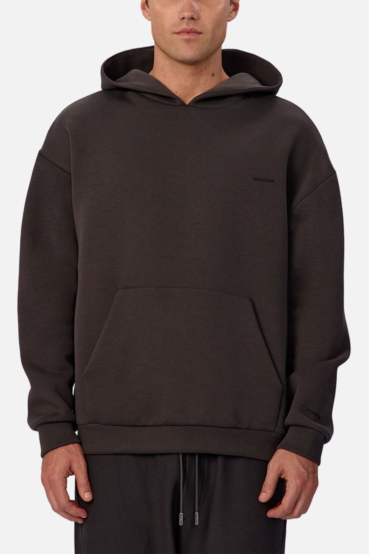 A person wears the Industrie The Tech Del Sur Hoodie—a loose, dark athletic hoodie with a front pocket and small left-chest logo—paired with drawstring pants, standing against a plain light background facing forward.