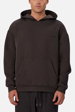 A person wears the Industrie The Tech Del Sur Hoodie—a loose, dark athletic hoodie with a front pocket and small left-chest logo—paired with drawstring pants, standing against a plain light background facing forward.