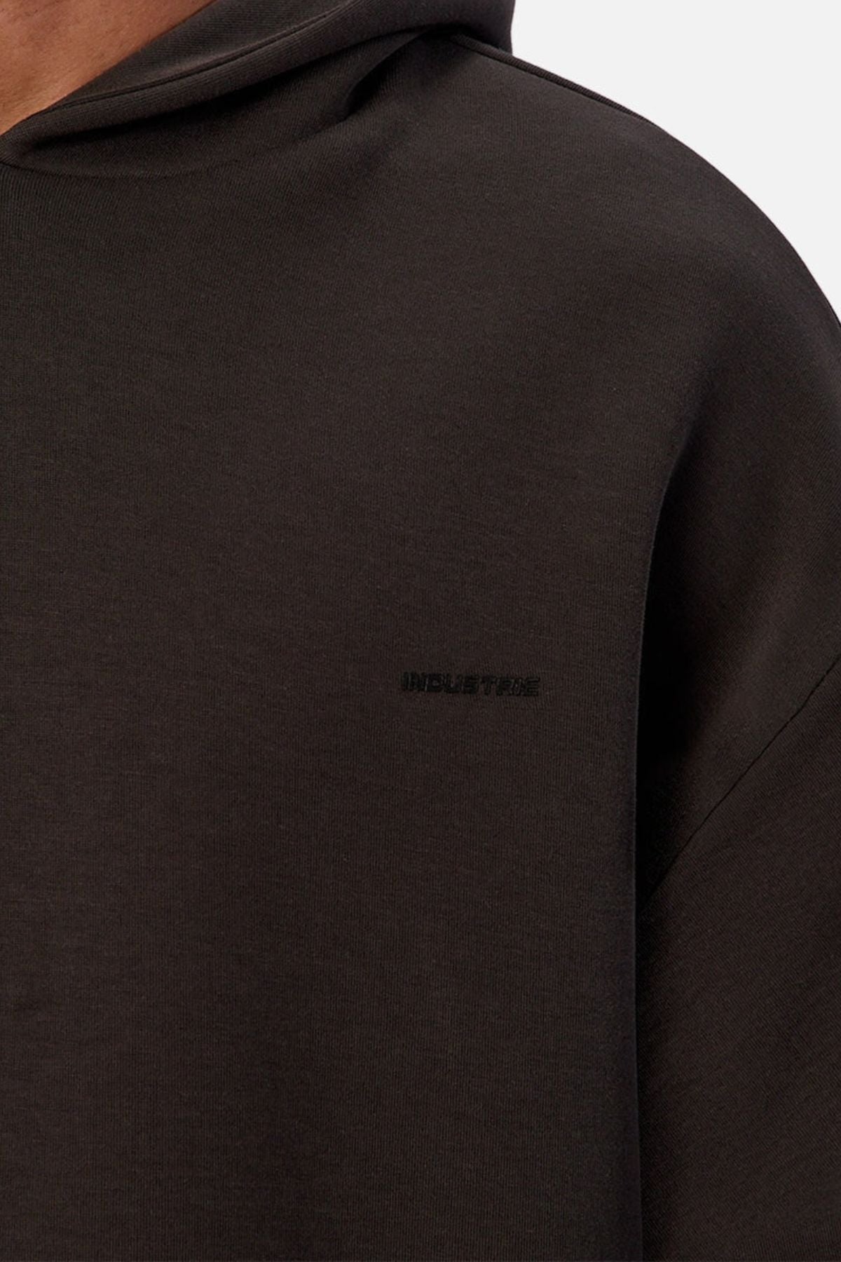 Close-up of the Industrie The Tech Del Sur Hoodie, a dark brown pullover featuring "INDUSTRIE" embroidered in small black text on the left chest. The soft fabric and partially seen hood reflect its athletic style.