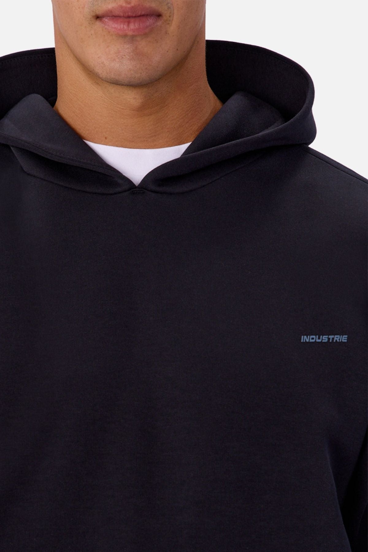 A close-up of a person in the Industrie The Tech Del Sur Hoodie, a black athletic pullover with a small "INDUSTRIE" logo on the left chest, layered over a white t-shirt. The person's face is partially visible.